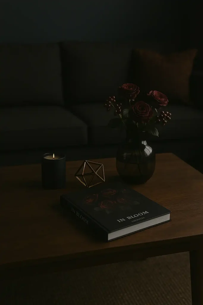 A dark walnut rectangular coffee table in a moody-toned living room with deep navy or charcoal walls. Decor includes a black candle, a gold or brass geometric sculpture, a single dark floral coffee table book, and a smoky glass vase with deep red roses or berries. Dim, ambient lighting.